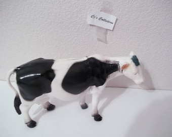 May include: Black and white plastic toy cow with a green and orange ear tag. The cow is standing on a white surface.  Cj's Collection.