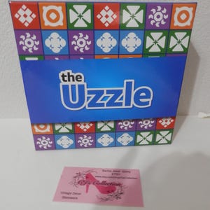 Può includere: Una scatola colorata con un motivo geometrico arancione, viola, verde e bianco. La scatola ha uno striscione blu con la scritta "the Uzzle". Un biglietto da visita rosa con un logo a tacco alto è di fronte alla scatola.