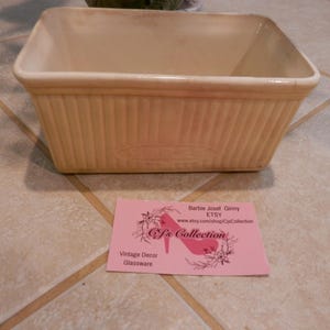 May include: An off-white ceramic baking dish, rectangular with ribbed sides. The dish has a flared shape and a smooth interior. A pink business card with text and a high-heeled shoe logo is in front of the dish. The card reads "J's Collection".