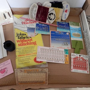 Puede incluir: Una caja llena de diversos artículos de costura, incluyendo carretes de hilo, agujas, ganchos y ojales, cinta de bies y un folleto titulado "Joins fabrics without stitching".