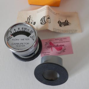 Puede incluir: Un comprobador de carburador vintage con un bisel cromado y una base negra. El medidor indica "Flow Meter" y "Carburetor Tester". La imagen incluye una tarjeta de visita rosa y un diagrama.