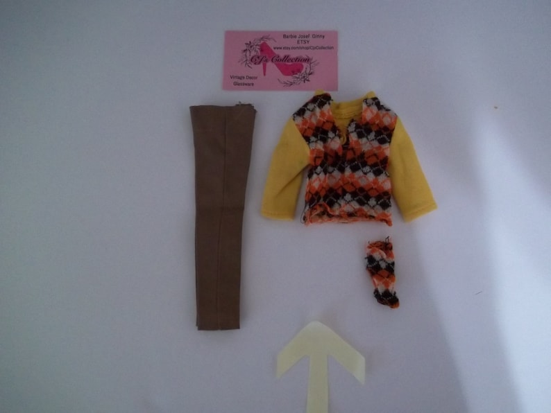 May include: A set of doll clothes including a brown pair of pants, a yellow sweater with a brown, orange, and white geometric pattern, and a pair of orange and brown patterned stockings.