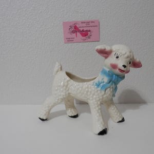 Könnte beinhalten: Ein Vintage-Pflanzgefäß in Form eines Lammes aus Keramik, weiß mit strukturierter Oberfläche. Das Lamm hat rosa Ohren und Wangen, eine blaue Schleife und schwarze Hufe. Der Pflanztopf ist für kleine Pflanzen oder Blumen gedacht.