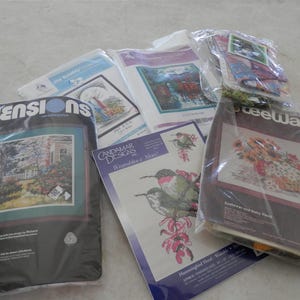 May include: A collection of cross-stitch patterns featuring various designs, including hummingbirds, a basket of flowers, and a spring scene. The patterns are packaged in plastic sleeves and include instructions and dimensions.