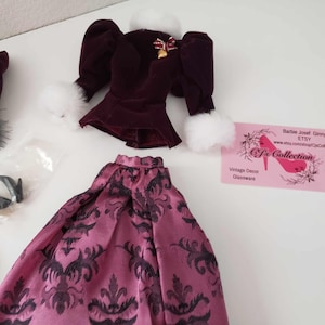 May include: A doll outfit set with a burgundy velvet jacket with white fur trim, a pink and black patterned skirt, a black hat with a feather, and a pair of black shoes. The outfit is for a Barbie doll.