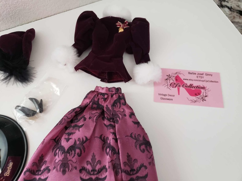 May include: A doll outfit set with a burgundy velvet jacket with white fur trim, a pink and black patterned skirt, a black hat with a feather, and a pair of black shoes. The outfit is for a Barbie doll.