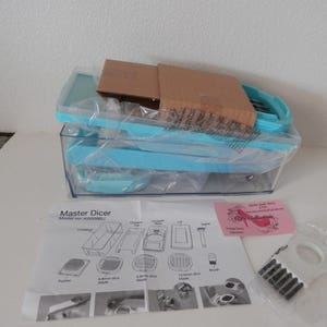 May include: A new Master Dicer set, featuring a clear container with a light blue frame and accessories. Includes a manual and additional components, all packaged in plastic. A pink card and a small white tool are also visible.