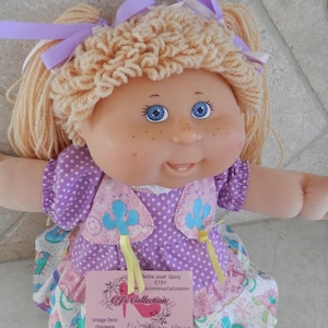 May include: A vintage Cabbage Patch Kid doll with blonde yarn hair and blue eyes. The doll wears a purple polka dot dress with a floral print skirt and a matching vest. The doll has a pink tag that says "CJ's Collection".
