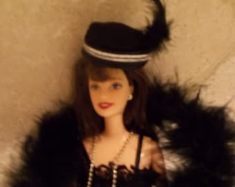May include: A black and white doll wearing a black hat with a feather, a black lace dress, and a pearl necklace. The doll is wearing a black feather boa.