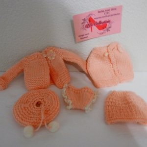 May include: A set of five knitted doll clothes in a peach color. The set includes a cardigan, a skirt, a dress, a hat, and a pair of booties.