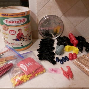 May include: A vintage TinkerToy construction set with 325 pieces. The set includes a cylindrical container, various plastic and wooden pieces in red, yellow, black, and blue, and a metal lid. The container features a child building a toy.