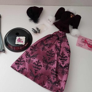 May include: A pink and black patterned dress with a black velvet jacket, a black hat, and a doll stand. The dress has a damask pattern and the jacket has a white fur trim. The doll stand is black and has a round base.