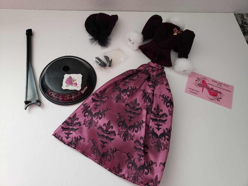 May include: A pink and black patterned dress with a black velvet jacket, a black hat, and a doll stand. The dress has a damask pattern and the jacket has a white fur trim. The doll stand is black and has a round base.