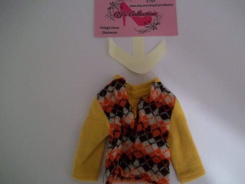 May include: A yellow long-sleeved shirt with a brown, orange, and white patterned vest. The vest has a diamond pattern.