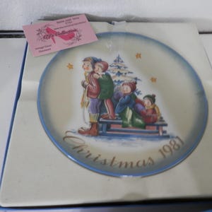 May include: A vintage Christmas plate from 1981, depicting children sledding in a winter scene. The plate has a blue border and gold lettering. The image shows children in winter clothing, a Christmas tree, and stars.