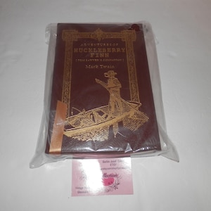 Può includere: Un libro con copertina rigida, "Adventures of Huckleberry Finn" di Mark Twain, con lettere dorate in rilievo e un'illustrazione di una barca. Il libro è sigillato in un sacchetto di plastica trasparente. La copertina è bordeaux.