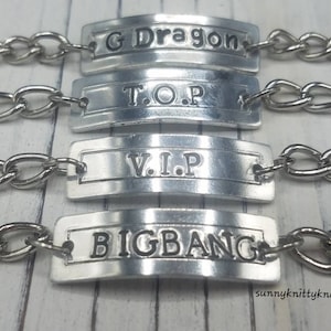 Könnte beinhalten: Vier silberne Armbänder mit Kettengliedern und rechteckigen Metalletiketten. Die Etiketten tragen die Worte "G Dragon", "T.O.P", "V.I.P" und "BIGBANG" eingraviert.