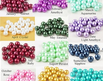 May include: A collection of colorful beads in various shades, including red, purple, green, white, and blue. Each pile of beads is labeled with a month and gemstone name, such as "January Garnet" and "February Amethyst."