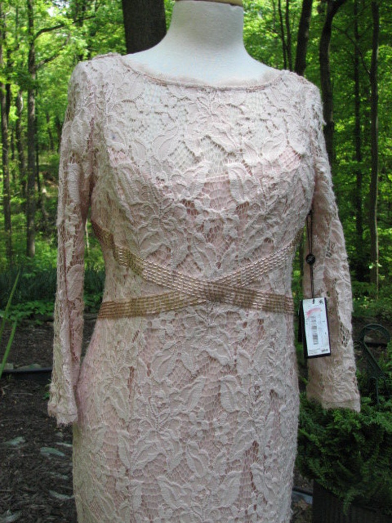 Pu&ograve; includere: Un abito in pizzo rosa cipria a maniche lunghe con una cintura di perline dorate. L'abito ha una scollatura rotonda e una silhouette aderente.