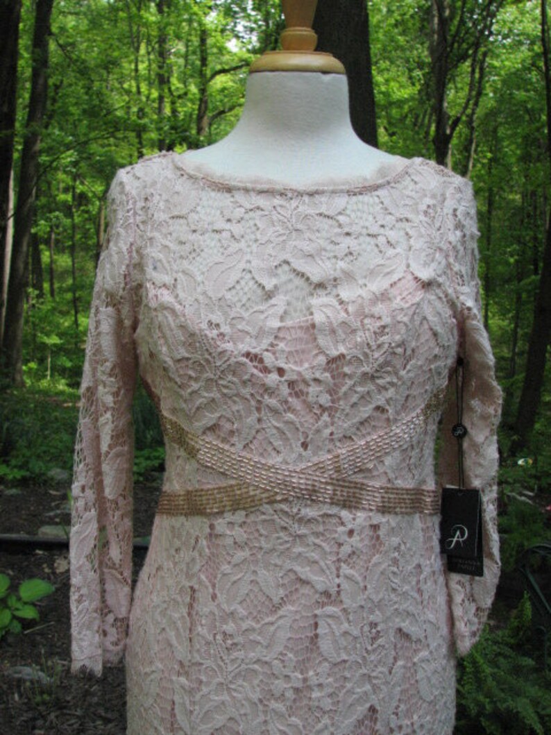 Pu&ograve; includere: Un abito in pizzo rosa cipria con una cintura di perline e maniche lunghe. L'abito ha una silhouette aderente e un delicato motivo floreale.