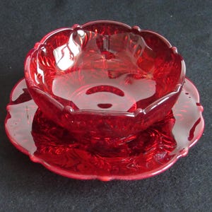 May include: A deep red glass bowl and matching saucer set featuring a translucent finish and a scalloped edge design. The glass surface displays a molded floral pattern with raised textures throughout the interior and exterior of the bowl and the plate.
