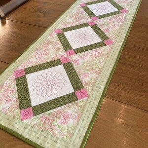 May include: A floral table runner with a light green border and a pink and white floral pattern. The runner features three square designs with pink flower embroidery, framed by green and pink accents. The runner is placed on a wooden table.