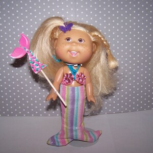 May include: A mermaid doll with blonde hair, freckles, and a smiling face. The doll wears a pink seashell top and a striped mermaid tail. It holds a pink and teal wand with a mermaid tail design. A purple butterfly decorates the doll's hair.