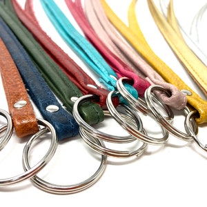 Könnte beinhalten: Sortiment an Leder-Schlüsselanhängern in verschiedenen Farben, darunter Schwarz, Braun, Grün, Blau, Rot, Rosa, Gelb und Silber. Jeder Schlüsselanhänger hat einen silbernen Metallring.