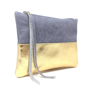 Puede incluir: Un peque&ntilde;o clutch de cuero gris y dorado con una borla gris larga. El clutch tiene un acabado texturizado sutil y el nombre de la marca "kaa" est&aacute; estampado en la parte delantera.