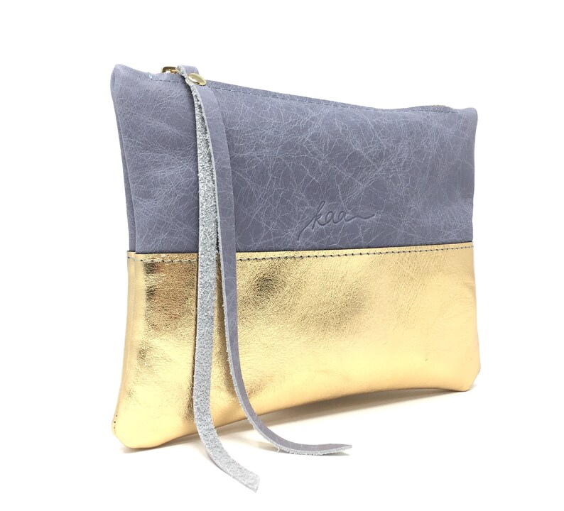 Puede incluir: Un peque&ntilde;o clutch de cuero gris y dorado con una borla gris larga. El clutch tiene un acabado texturizado sutil y el nombre de la marca "kaa" est&aacute; estampado en la parte delantera.