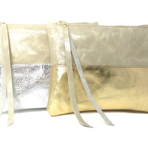 Puede incluir: Tres clutches de cuero con cierre de cremallera. Los clutches son de dos tonos, con la mitad superior en beige claro y la mitad inferior en plata, oro y plata. Cada clutch tiene una correa larga de cuero.