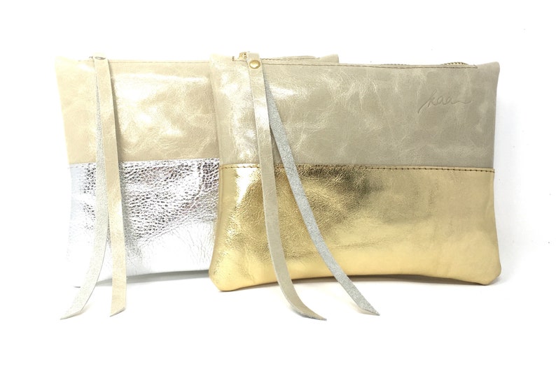 Puede incluir: Tres clutches de cuero con cierre de cremallera. Los clutches son de dos tonos, con la mitad superior en beige claro y la mitad inferior en plata, oro y plata. Cada clutch tiene una correa larga de cuero.