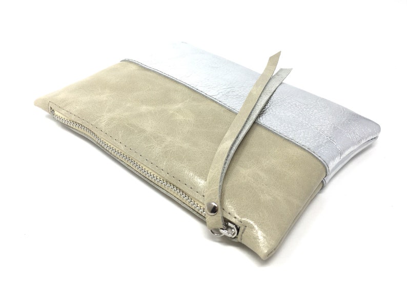 Puede incluir: Un clutch de cuero beige y plateado con cierre de cremallera y una correa larga.