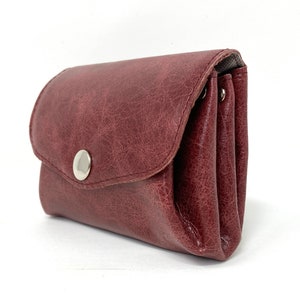 May include: A burgundy leather wallet with a silver button closure. The wallet is folded and has a textured surface.