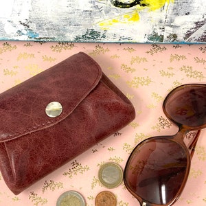 May include: A brown leather wallet with a snap closure and a silver button. The wallet is on a pink background with gold accents. There are also sunglasses and coins in the image.
