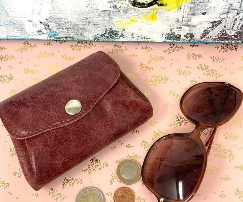 May include: A brown leather wallet with a snap closure and a silver button. The wallet is on a pink background with gold accents. There are also sunglasses and coins in the image.