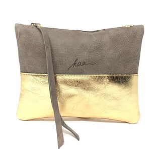 Puede incluir: Un clutch de cuero gris y dorado con cierre de cremallera y una correa larga. El clutch tiene la palabra "kaa" grabada en la parte delantera.