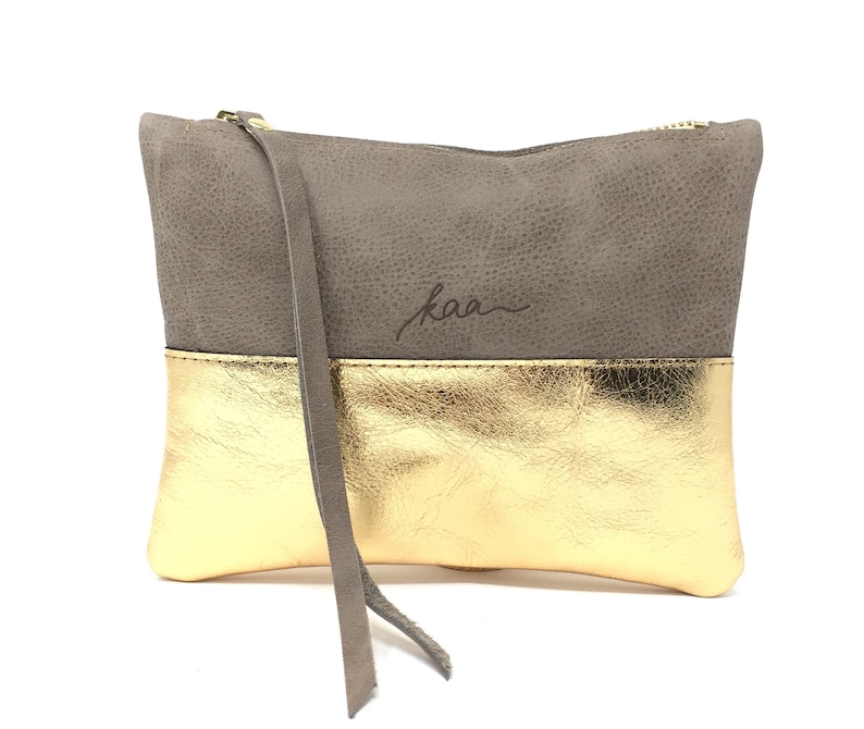 Puede incluir: Un clutch de cuero gris y dorado con cierre de cremallera y una correa larga. El clutch tiene la palabra "kaa" grabada en la parte delantera.