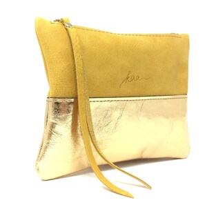 Puede incluir: Un peque&ntilde;o clutch de cuero amarillo y dorado con una correa larga. El clutch tiene la palabra "kaa" grabada en la parte delantera.