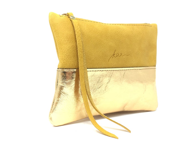 Puede incluir: Un peque&ntilde;o clutch de cuero amarillo y dorado con una correa larga. El clutch tiene la palabra "kaa" grabada en la parte delantera.