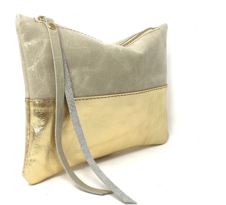 Puede incluir: Un peque&ntilde;o bolso de mano rectangular hecho de cuero. La mitad superior del bolso es de color gris claro y la mitad inferior es de color dorado. El bolso tiene una cremallera y una correa larga y fina.