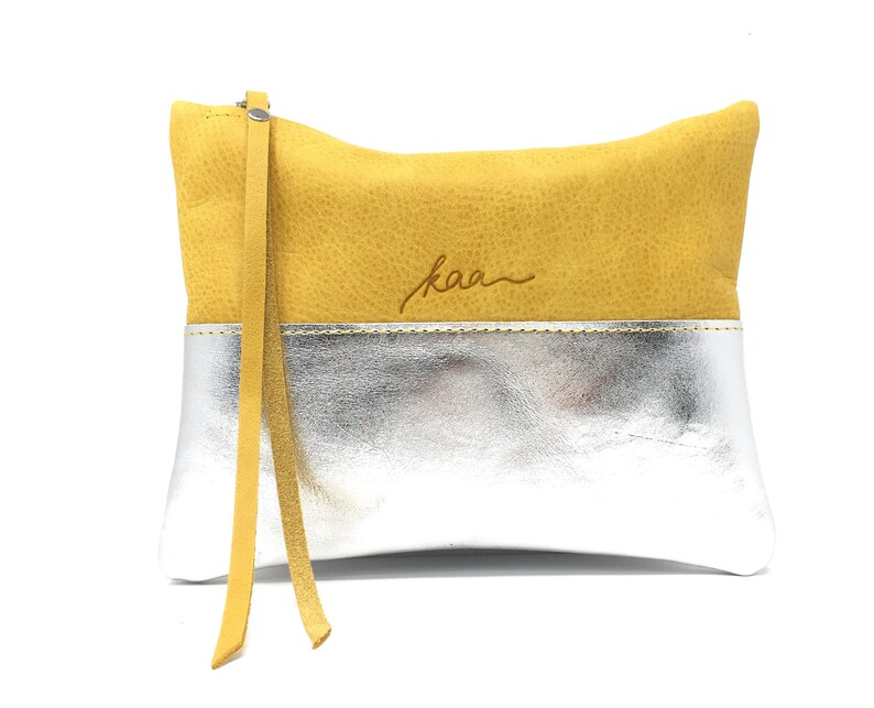 Puede incluir: Un clutch de cuero amarillo y plateado con una correa de cuero marr&oacute;n y la palabra "kaa" grabada en la parte delantera.