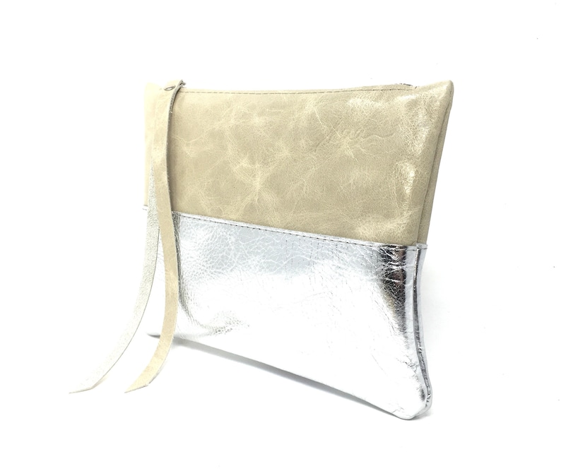 Puede incluir: Un clutch de cuero beige y plateado con una correa larga beige. El clutch tiene una cremallera.