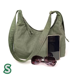 May include: A green canvas shoulder bag with a long strap and a zip closure. The bag is sitting on a white surface with a pair of sunglasses and a black smartphone.