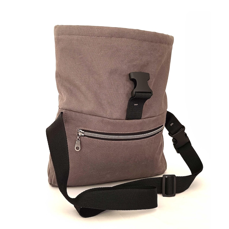 May include: A gray canvas messenger bag with a black adjustable strap and buckle closure. The bag features a zippered front pocket and a flap closure with a black buckle. The bag is designed to be worn over the shoulder.
