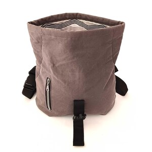 May include: A dusty rose-colored backpack with a black strap and buckle closure. The bag has a zippered pocket on the front and a striped interior lining. The bag is made of a soft, textured material.