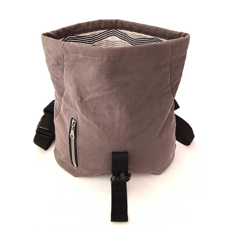 May include: A dusty rose-colored backpack with a black strap and buckle closure. The bag has a zippered pocket on the front and a striped interior lining. The bag is made of a soft, textured material.