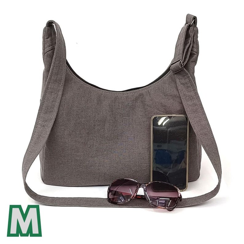May include: A grey canvas shoulder bag with a long strap. The bag is open and a pair of sunglasses and a mobile phone are inside.
