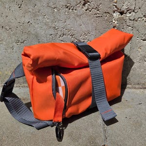 May include: Bright orange messenger bag with a gray adjustable strap and black buckle. The bag has a zippered pocket with a checkered pattern inside. The bag is made of a durable material and is designed for carrying items.