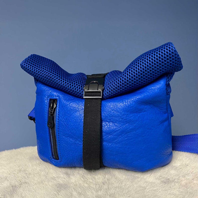 May include: A vibrant blue bag with a textured exterior and a mesh top. The bag features a black strap with a buckle closure and a black zipper. The bag is set against a blue background.